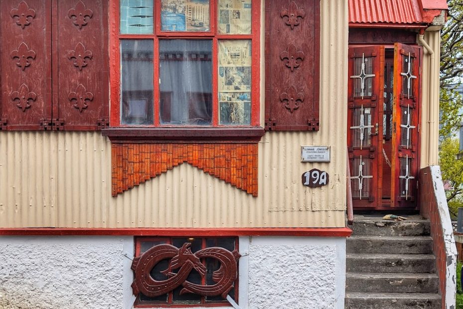 Facade of a home in Reykjavik Iceland. There is a window covered in newspaper in the foreground. The shutters have fleur-de-lis on them. There is a smaller basement window decorated with a carved dragon. A small stairway and a wooden door that is partially open sits to the right of the window and is slightly recessed.