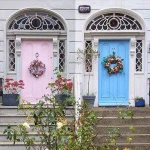 Two vibrantly colored doors, one pink and the other blue, stand side-by-side on a sunny day, each adorned with a wreath and framed by ornate architectural details on a white building.