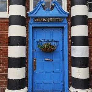 Vibrant blue wooden door with a hanging flower basket, framed by bold black and white striped columns in Buffalo, New York.