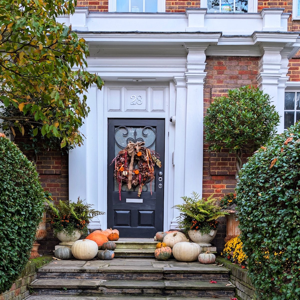 Elegant black London townhouse doorway with an autumn door wreath and steps covered in colorful heirloom pumpkins and seasonal greenery.