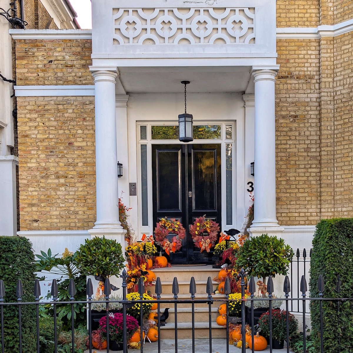 Grand white London townhouse entryway with a black front door, flanked by stone steps lined with autumn leaves and colorful pumpkins.