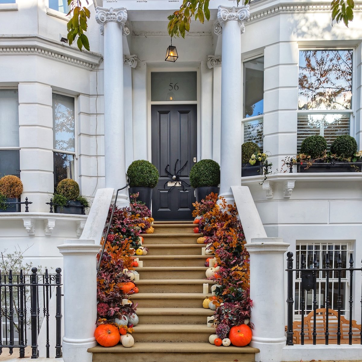 Black double front doors on a brick London townhouse featuring fall wreaths, potted chrysanthemums, and orange pumpkins along the porch steps.
